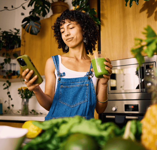 young woman in kitchen with green smoothie and phone